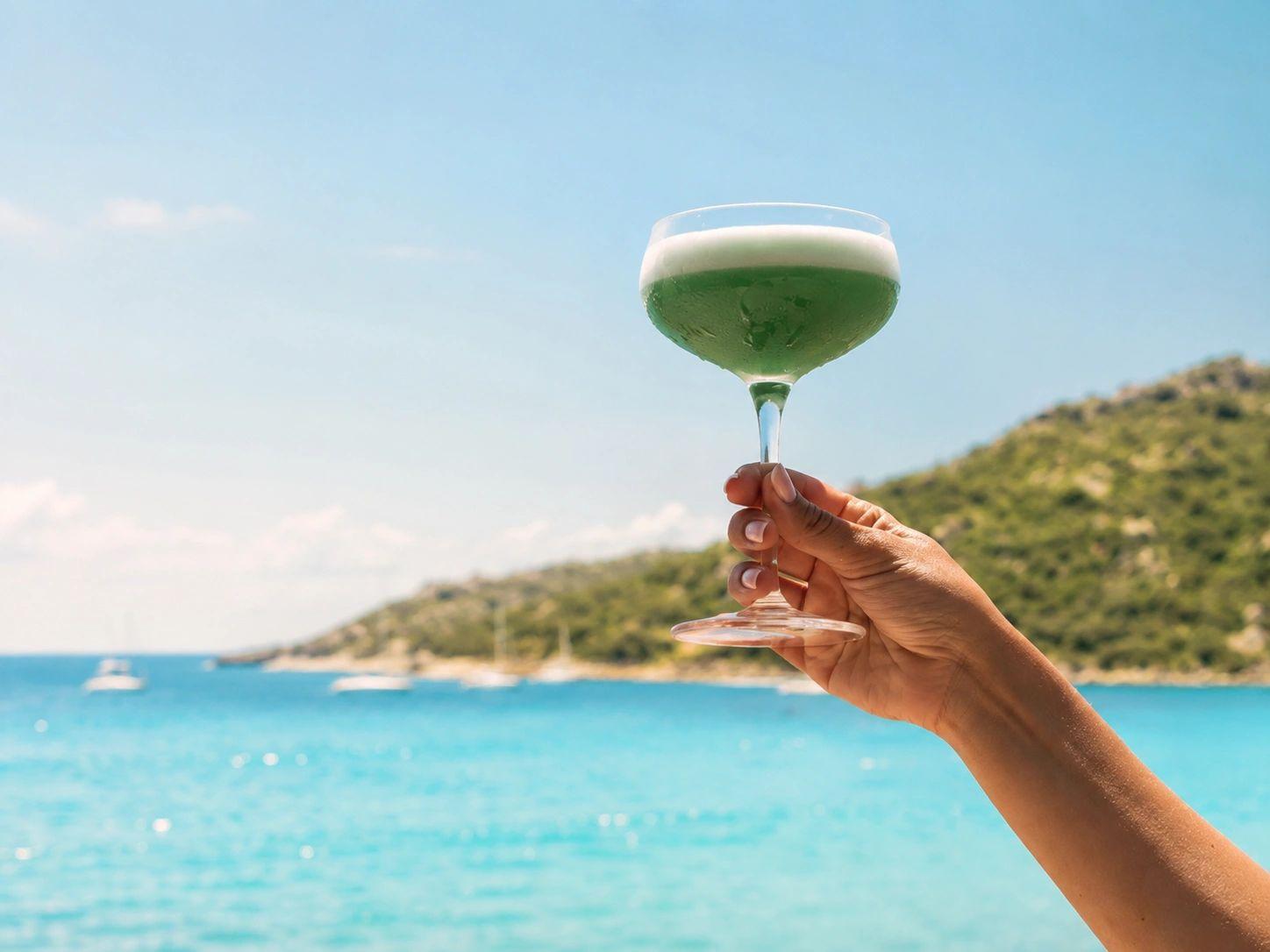 Une main tient un verre de cocktail vert face à la mer turquoise de Santa Giulia